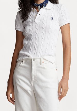 Femme portant un polo blanc à manches courtes en maille torsadée, un pantalon blanc taille haute et un foulard bleu à motifs autour du cou.