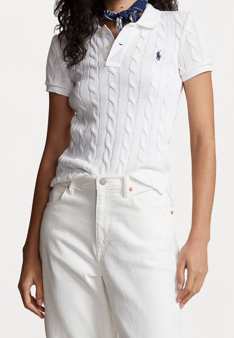 Femme portant un polo blanc à manches courtes en maille torsadée, un pantalon blanc taille haute et un foulard bleu à motifs autour du cou.