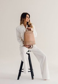 Světle růžový kožený batoh zaobleného tvaru, s jemným detailem loga a nastavitelnými popruhy, umístěný na neutrálním pozadí.