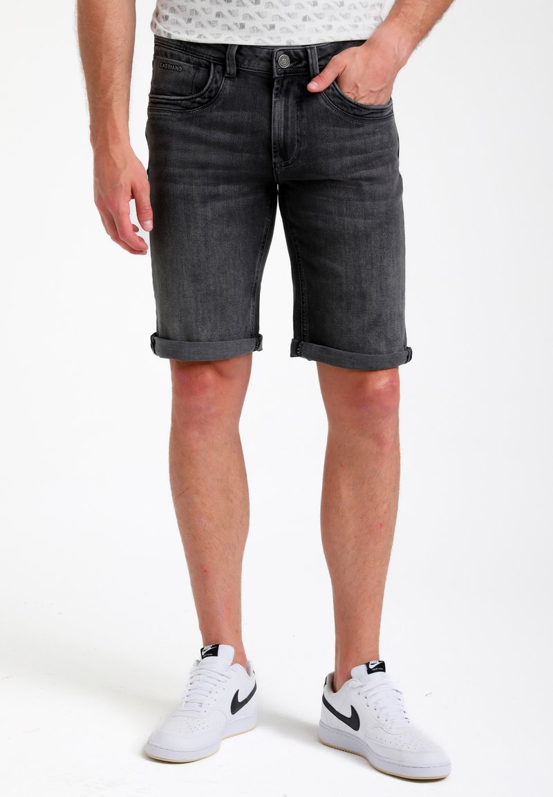 Pantalones cortos de mezclilla negra con dobladillos enrollados, que cuentan con cinco bolsillos y un cierre frontal con botón. Combinados con zapatillas blancas con un diseño de swoosh negro.
