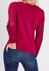 Karmesinroter Pullover mit einfarbigem Körper und strukturierten Spitzenärmeln, runder Ausschnitt und gerippte Bündchen. Getragen mit dunkelblauen Jeans.