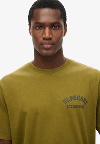 Zielona bawełniana koszulka z krótkim rękawem i okrągłym dekoltem. Na przedniej stronie znajduje się niebieski nadruk z napisem "Superdry Los Angeles". Miękka struktura.