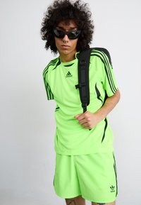 Neonas žalias sportinis marškinėliai ir šortai su juodais akcentais, su Adidas logotipu. Modelis dėvi juodus akinius nuo saulės ir juodą kuprinę.