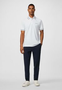Biely polo tričko s námorníckymi akcentmi a logom, skombinované s tmavomodrými nohavicami a krémovými teniskami, prezentované na modeli na bezfarebnom pozadí.