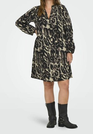 Kvinde iført en løs kjole med leopardprint, lange ærmer og sorte mellemlange støvler, stående med den ene hånd på hoften.