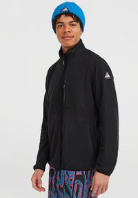 Giacca in pile nera con tasca con zip, colletto rialzato e accenti neri. Una cuffia in maglia blu con logo è indossata sulla testa.