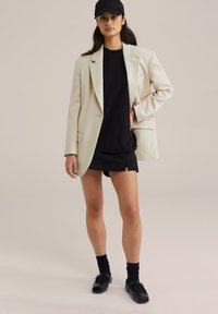 Béžový blazer přes černé tričko a černé šortky, doplněný černými ponožkami a botami. Celý outfit dotváří černá čepice.