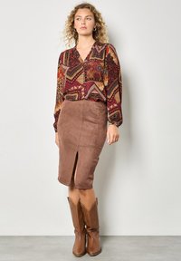 Brauner Wildledrock mit einem Schlitz vorne, kombiniert mit einer gemusterten weinrotfarbenen Bluse mit geometrischen Designs. Das Model trägt braune Cowboystiefel.