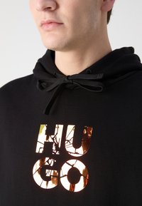 Sudadera negra con un acabado texturizado, que presenta una gran impresión de foil dorado agrietado con la palabra "HUGO" en el frontal. Cierre con cordones en el cuello.