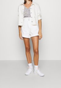 Femme portant une veste en denim blanche et un short taille haute blanc sur un haut à fleurs violettes, accompagnée de chaussettes blanches et de baskets blanches.