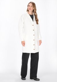 Biały sweterek dzianinowy z zygzakowatą fakturą, z drewnianymi guzikami z przodu. Połączony z czarnymi spodniami o szerokich nogawkach i botkami za kostkę.