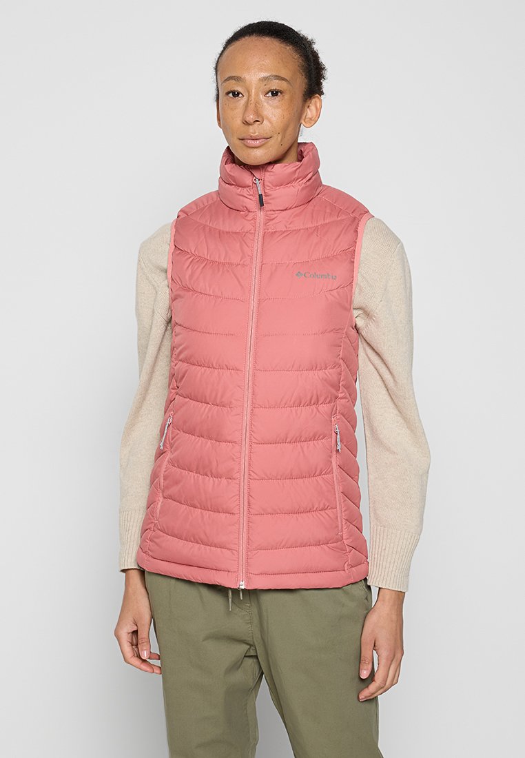 Modella che indossa un giubbotto smanicato trapuntato rosa, chiuso con la zip sopra un maglione beige e pantaloni verde oliva, in piedi di fronte a uno sfondo neutro.