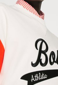 Felpa di colore crema con accenti rossi, dotata di un logo ricamato in nero con la scritta "Bold" e la parola "Athletic", indossata su un colletto con righe rosse e bianche.