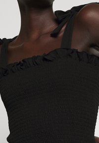 Musta smocked-top, jossa on röyhelöreunat ja solmittavat hihnat. Kangas on joustavaa ja tekstuuriltaan rönsyilevää, luoden tiukasti istuvan ilmeen.