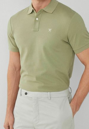 Olivgrünes Polohemd mit geripptem Kragen, zwei Knöpfen und gesticktem Logo. Begleitet von hellgrauen Hosen.