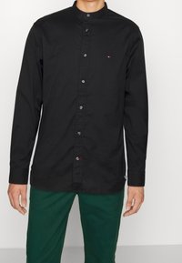 Camiseta negra de manga larga con cuello alto, cierre de botones y un pequeño logo en el pecho. La tela es suave con un ligero brillo.