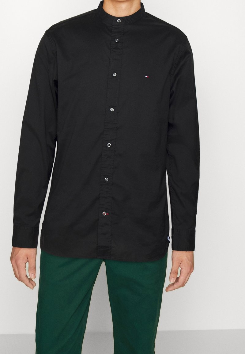 Camiseta negra de manga larga con cuello alto, cierre de botones y un pequeño logo en el pecho. La tela es suave con un ligero brillo.