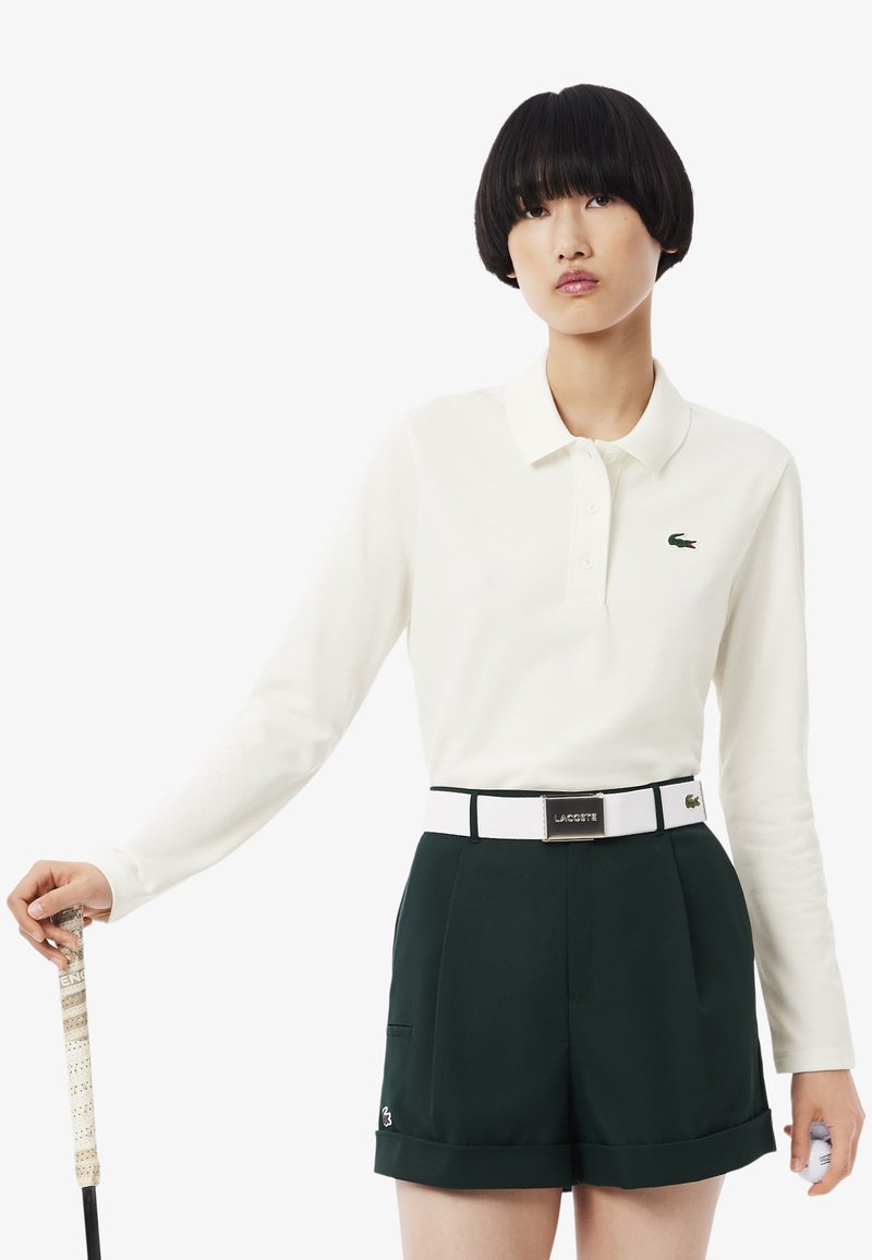 Weißes Langarm-Poloshirt mit grünem Krokodil-Logo, kombiniert mit dunkelgrünen Shorts, die einen weißen Gürtel und dezente Markendetails aufweisen.