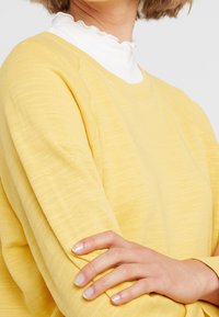 Maglione giallo con un motivo a maglia testurizzata, caratterizzato da un collo rotondo e spalle scese, indossato sopra una camicia bianca con colletto arricciato.