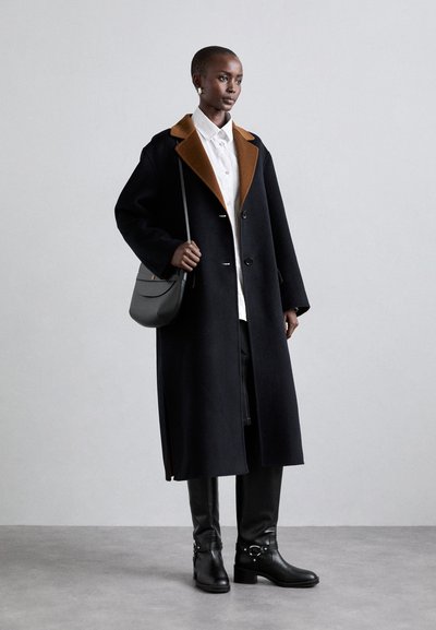 Langer schwarzer Mantel mit braunem Kragen, schwarze enganliegende Hose und Stiefeletten. Das Modell hält eine graue Tasche und steht vor einem hellen Hintergrund.