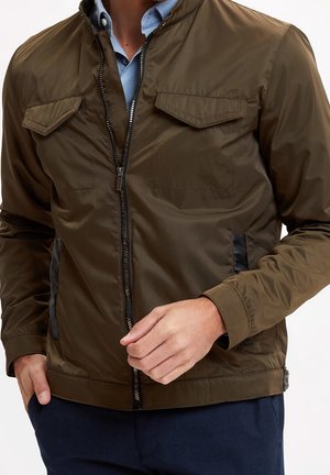 Mann trägt eine olivgrüne Jacke mit Reißverschluss und Klappentaschen auf der Brust über einem blauen Hemd und richtet die Jacke mit einer Hand in der Taillenregion.