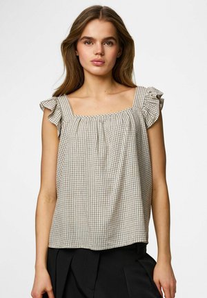 Giovane donna con top beige senza maniche con motivo a griglia nero e spalline arricciate, abbinato a pantaloni neri, su sfondo semplice.