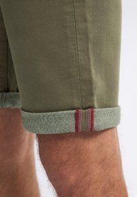 Shorts vert olive avec des ourlets retroussés, présentant une bande texturée vert clair et des accents de surpiqûres rouges sur l'ourlet et l'ourlet retroussé.