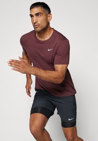 Nike kortermet skjorte i burgunder og svarte shorts med grå detaljer; skjorten har rund hals, shortsen er knelang med elastikk i kanten.