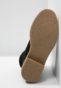 Bottine noire en daim avec une semelle en caoutchouc texturée ; taille 37 indiquée sur la semelle, inclinée contre un fond blanc.