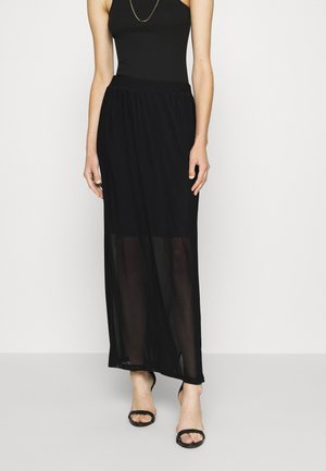 Kobieta ubrana w czarny bezrękawnik, przejrzystą czarną długą spódnicę, czarne sandały na otwartym palcu oraz cienki złoty naszyjnik, stojąca na białym tle.