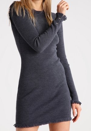 Mujer con un vestido ajustado de manga larga en punto acanalado gris oscuro, con bordes desflecados y detalles de botones en los puños.