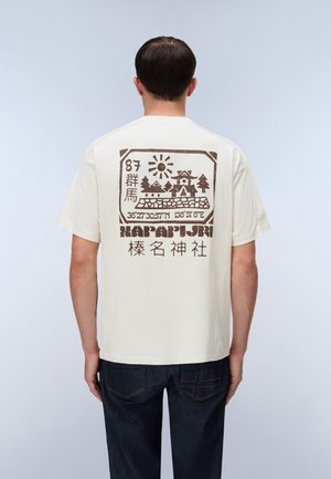 Homme portant un t-shirt crème avec un motif brun de soleil, maisons, arbres et texte japonais dans le dos, assorti à un jean bleu foncé.