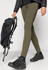 Legíny olivovo zelenej farby z elastického materiálu, s hladkou textúrou a priliehavým strihom, doplnené čiernymi turistickými topánkami a čiernym batohom.