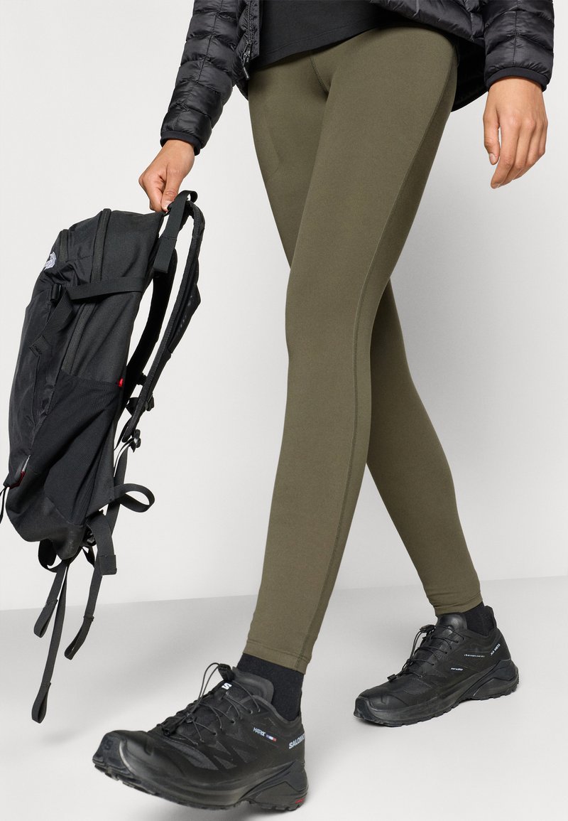 Leggings de color verde oliva hechos de material elástico, con una textura suave y un diseño ajustado, combinados con zapatos de senderismo negros y una mochila negra.