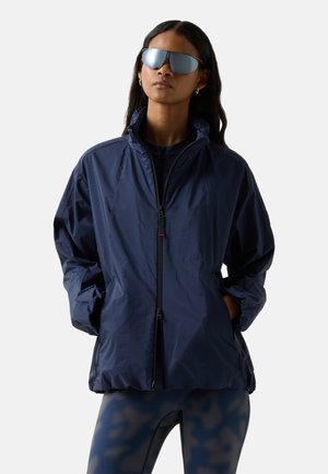 Frau trägt übergroße marineblaue Jacke mit Reißverschluss, Leggings mit Tarnmuster und große reflektierende Sonnenbrille, steht mit den Händen in den Taschen.