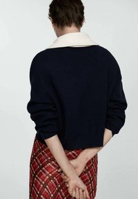 Pull bleu marine avec col blanc, design court et manches volumineuses ; associé à une jupe à carreaux rouge. Vue de dos montrant les mains jointes.