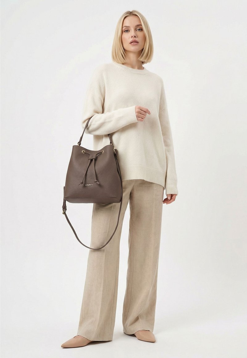 Femme aux cheveux blonds portant un pull crème et un pantalon beige, tenant un sac seau en cuir marron foncé avec une fermeture à cordon.