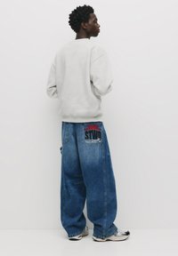 Hanorac supradimensionat de culoare gri deschis, purtat cu pantaloni din denim albastru largi, având o etichetă logo roșie și neagră pe buzunarul din spate. Adidași alb-negru.