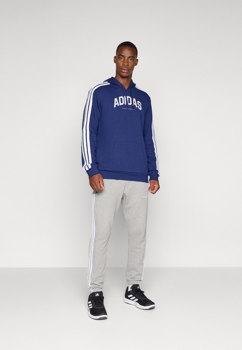 Marineblauwe hoodie met witte "ADIDAS" logo, grijze joggingbroek met witte zijstrepen en zwarte sportschoenen, alles gepresenteerd tegen een neutrale achtergrond.