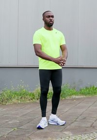 Neonowożółta koszulka sportowa, czarne legginsy kompresyjne oraz białe buty do biegania z niebieskimi akcentami. Pozycja stojąca na chodniku z trawą.