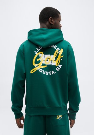 Mann trägt grünen Hoodie und passende Hose mit gelbem und weißem "Golf Augusta, GA"-Logo auf der Rückseite und Tasche, gesehen von hinten.