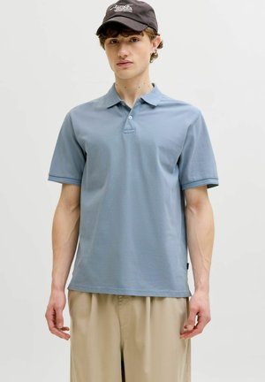 Fiatal férfi világoskék pólóingben, bézs nadrágban és fekete sapkában, amelyen fehér logó látható, egy egyszínű világos háttér előtt áll.