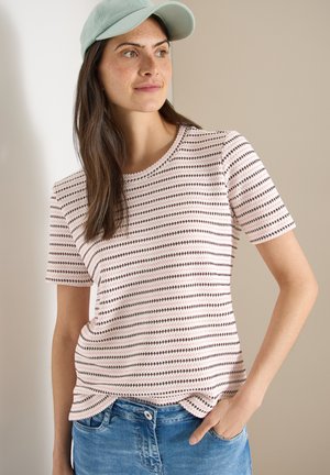 Frau mit langen braunen Haaren, die ein gestreiftes kurzärmeliges Shirt, eine hellgrüne Kappe und blaue Jeans trägt, mit einer Hand in der Tasche, die seitlich schaut.