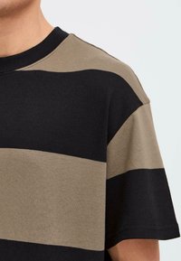 Le T-shirt à manches courtes présente des rayures horizontales noires et kaki, une texture douce, et un col rond. Design minimaliste avec des couleurs contrastantes.