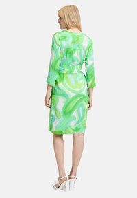 Grünes Kleid mit 3/4-Ärmeln, das ein wirbelndes Muster in verschiedenen Grüntönen und Weiß zeigt. Der Stoff wirkt leicht; tailliert mit einem Gürtel.