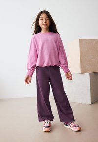 Růžová mikina s dlouhým rukávem a manžetami, v kombinaci s volnými fialovými kalhotami. Barevné tenisky s vzorovaným designem doplňují outfit.