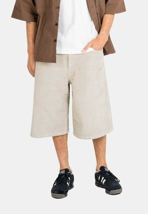 Man wearing beige knee-length corduroy shorts, white t-shirt, brown short-sleeve shirt, and black sneakers with white stripes, one hand in pocket.