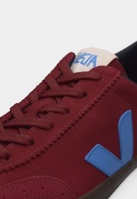 Gros plan sur une basket bordeaux avec des lacets assortis et un logo "V" bleu sur le côté, avec une languette beige arborant un texte bleu.