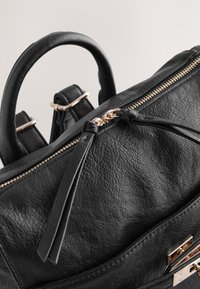 Zwarte leren rugzak met een textuurachtige oppervlak, gouden ritssluiting, bovenkanten handgrepen en decoratieve knoopdetail aan de ritstrekker.