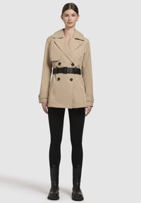 Beige dubbelzijdige trenchcoat met een zwarte riem, voorzien van brede revers en knoopdetails, gecombineerd met zwarte leggings en enkellaarsjes.